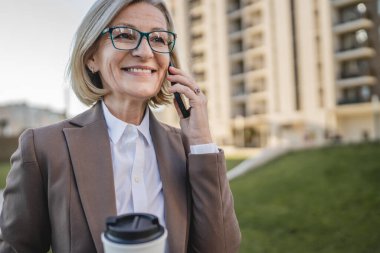 Olgun bir kadın binaların önünde cep telefonuyla konuşuyor ve elinde kahve, modern iş ve yaşam tarzı konsepti tutuyor.