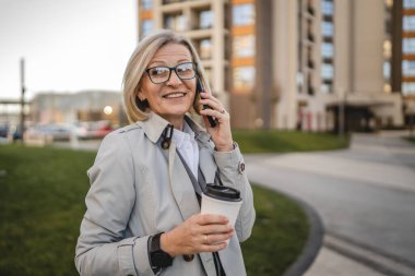 Olgun bir kadın binaların önünde cep telefonuyla konuşuyor ve elinde kahve, modern iş ve yaşam tarzı konsepti tutuyor.