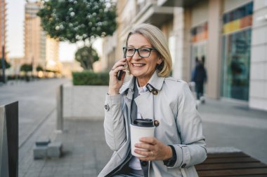 Olgun bir kadın binaların önünde cep telefonuyla konuşuyor ve elinde kahve, modern iş ve yaşam tarzı konsepti tutuyor.