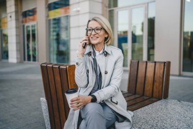Olgun bir kadın binaların önünde cep telefonuyla konuşuyor ve elinde kahve, modern iş ve yaşam tarzı konsepti tutuyor.