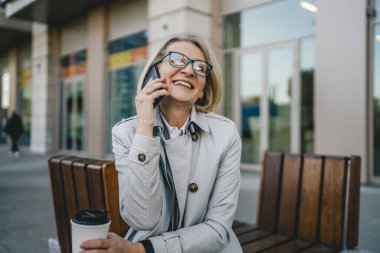 Olgun bir kadın binaların önünde cep telefonuyla konuşuyor ve elinde kahve, modern iş ve yaşam tarzı konsepti tutuyor.