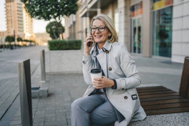 Olgun bir kadın binaların önünde cep telefonuyla konuşuyor ve elinde kahve, modern iş ve yaşam tarzı konsepti tutuyor.