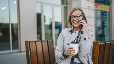 Olgun bir kadın binaların önünde cep telefonuyla konuşuyor ve elinde kahve, modern iş ve yaşam tarzı konsepti tutuyor.