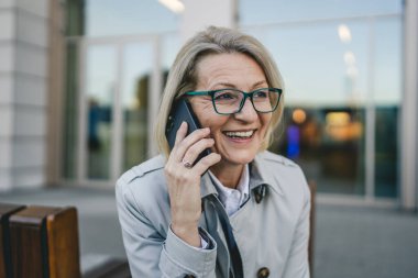 Binaların önünde cep telefonuyla konuşan olgun bir kadın, modern iş ve yaşam tarzı kavramı.