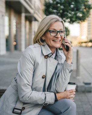 Olgun bir kadın binaların önünde cep telefonuyla konuşuyor ve elinde kahve, modern iş ve yaşam tarzı konsepti tutuyor.