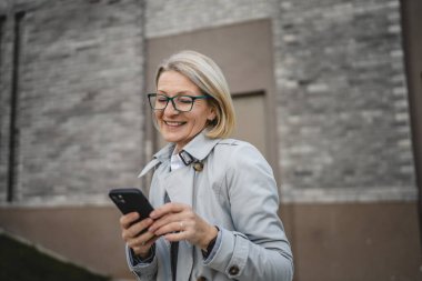 Modern bir kadın şehirde yetişkin bir kadın cep telefonu kullanıyor kısa mesajlar yolluyor ya da internete takılıyor gözlüklü sarışın bir kadın.