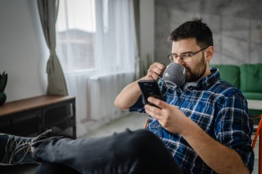 Yetişkin adam ev gezintisinde cep telefonu akıllı telefonu kullanır ya da bir fincan çay için kısa mesajlar gönderir.