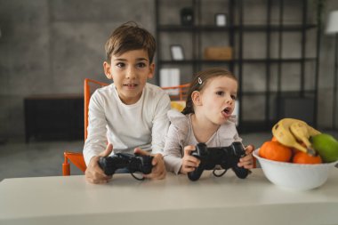 İki çocuk, küçük beyaz erkek ve kız kardeş mutlu çocuklar erkek ve kız kardeşler oyun konsolu oyunu oynarken evde otururken oyun konsolu ya da kumanda ile gerçek insanlar gerçek aile eğlencesi konsepti