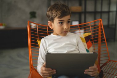 Bir beyaz çocuk evde oturup dijital tablet kullanıyor.