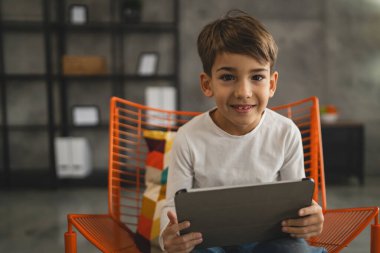 Bir beyaz çocuk evde oturup dijital tablet kullanıyor.
