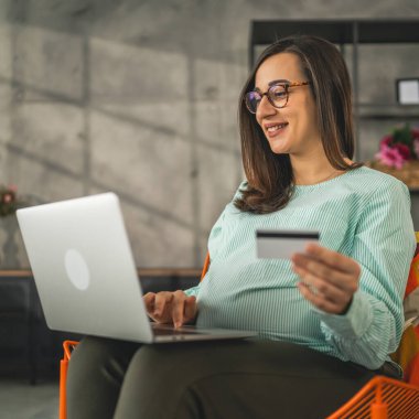 Hamile bir kadın online alışveriş yapıyor. Kredi kartı tutuyor. Evde oturuyor.