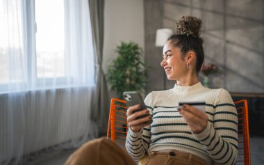 Yetişkin beyaz bir kadın mutlu bir şekilde gülümseyerek evde oturuyor banka kredi kartlarını online alışveriş konseptinde tutuyor gerçek insanlar uzay e-ticaretini kopyalıyor ya da rezervasyon yaptırıyor.