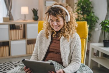 Kıvırcık saçlı beyaz bir kadın evde oturup dijital tablet ve kulaklık kullanarak film ya da dizi izliyor ya da mutlu bir gülümseme videosu çekiyor.
