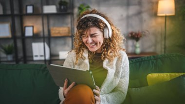 Kıvırcık saçlı beyaz bir kadın evde oturup dijital tablet ve kulaklık kullanarak film ya da dizi izliyor ya da mutlu bir gülümseme videosu çekiyor.