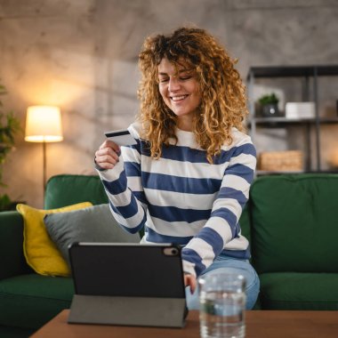 Evde mutlu gülümseyen yetişkin bir kadın banka kartı ve dijital tablet alışverişi e-bankacılık konseptini tutuyor. Gerçek insanlar uzay e-ticaretini kopyalıyor ya da rezervasyon yaptırıyor.