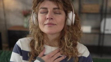 Yetişkin bir kadın Kafkas kadın milenyumu kulaklıkları online rehberli meditasyon için kullanıyor evde gözleri kapalı farkındalık yogası yapıyor gerçek insanlar özbakım kavramı yavaş çekim
