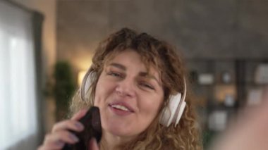 Kafkasyalı, kulaklıklı yetişkin bir kadın evde cep telefonu ile müzik dinliyor. Mutlu bir gülümseme. Keyfinize bakın.