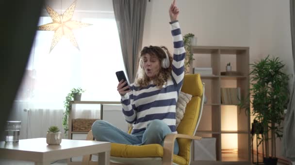 一个拿着耳机的高加索女人在家里用手机听音乐 网上开心地笑着唱着快乐独享一个复制的空间 — 图库视频影像