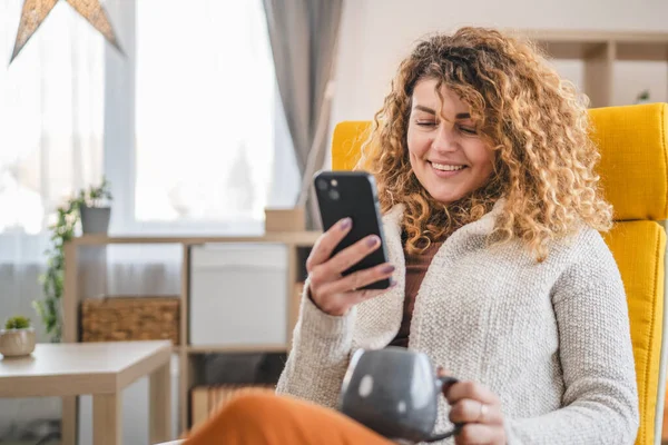 自宅でカーリーヘアを持つ1人の女性は 携帯電話のスマートフォンのSmsテキストを使用したり インターネットのオンラインソーシャルネットワークを閲覧します — ストック写真