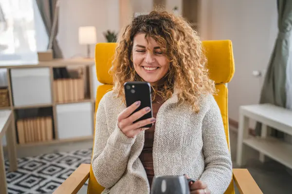 自宅でカーリーヘアを持つ1人の女性は 携帯電話のスマートフォンのSmsテキストを使用したり インターネットのオンラインソーシャルネットワークを閲覧します — ストック写真
