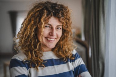 Evde kıvırcık saçlı, mutlu gülümseyen yetişkin beyaz kadın portresi.