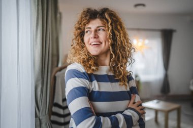 Evde kıvırcık saçlı, mutlu gülümseyen yetişkin beyaz kadın portresi.