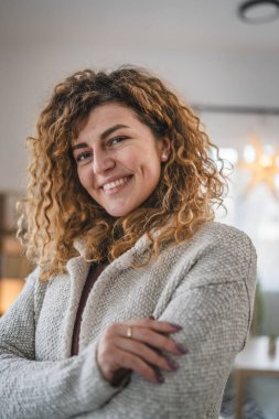 Evde kıvırcık saçlı, mutlu gülümseyen yetişkin beyaz kadın portresi.