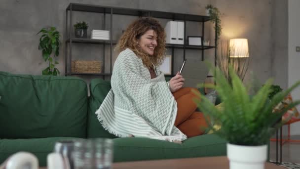 一个头发卷曲的高加索女人坐在家里用数码平板电脑看电影或在线连续剧 或者有视频叫快乐微笑 享受慢动作的快乐时光 — 图库视频影像
