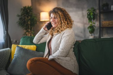 Evde kıvırcık saçlı beyaz bir kadın var. Cep telefonuyla telefon ediyor. Mutlu bir gülümseme.