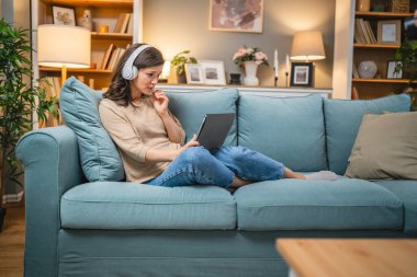 Yetişkin bir kadın beyaz kadın evde film videosu izlerken kanepeye oturur dijital tablet kullanır ya da video görüşmesi yapar.