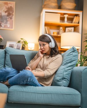 Yetişkin bir kadın beyaz kadın evde film videosu izlerken kanepeye oturur dijital tablet kullanır ya da video görüşmesi yapar.