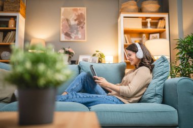 Yetişkin bir kadın beyaz kadın evde film videosu izlerken kanepeye oturur dijital tablet kullanır ya da video görüşmesi yapar.