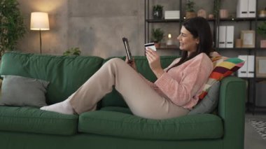 Genç bir yetişkin kadın evde oturup kredi kartı alışverişini internetten yapıyor.
