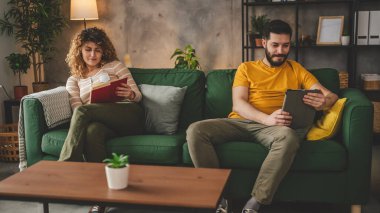 Erkek ve kadın Kafkasyalı yetişkin çift kitap okuyor ve evlerinde kanepe yatağında dijital tablet kullanıyorlar.