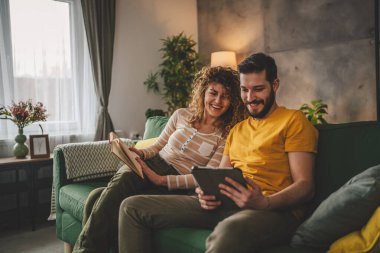 Erkek ve kadın Kafkasyalı yetişkin çift kitap okuyor ve evlerinde kanepe yatağında dijital tablet kullanıyorlar.