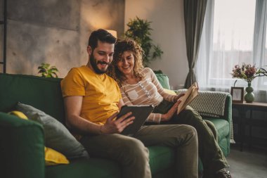 Erkek ve kadın Kafkasyalı yetişkin çift kitap okuyor ve evlerinde kanepe yatağında dijital tablet kullanıyorlar.