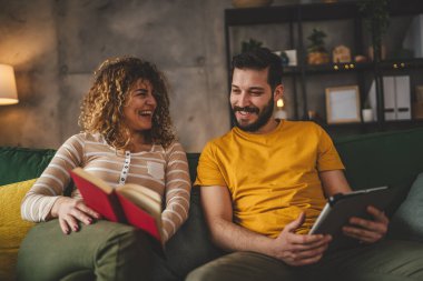 Erkek ve kadın Kafkasyalı yetişkin çift kitap okuyor ve evlerinde kanepe yatağında dijital tablet kullanıyorlar.