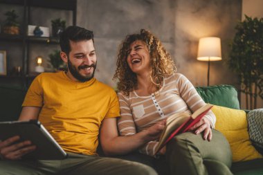 Erkek ve kadın Kafkasyalı yetişkin çift kitap okuyor ve evlerinde kanepe yatağında dijital tablet kullanıyorlar.