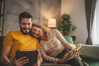 Erkek ve kadın Kafkasyalı yetişkin çift kitap okuyor ve evlerinde kanepe yatağında dijital tablet kullanıyorlar.