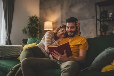 Erkek ve kadın Kafkasyalı yetişkin çift evde kanepe yatağında kitap okuyor.