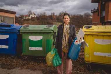 Bir yetişkin kadın cam, kağıt ve plastik sıfır atık konsepti için geri dönüşüm kutularının önünde yeşil sarı ve mavi poşetlerle duruyor.