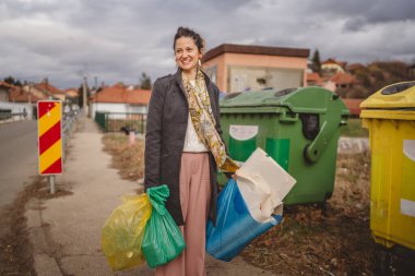 Bir yetişkin kadın cam, kağıt ve plastik sıfır atık konsepti için geri dönüşüm kutularının önünde yeşil sarı ve mavi poşetlerle duruyor.