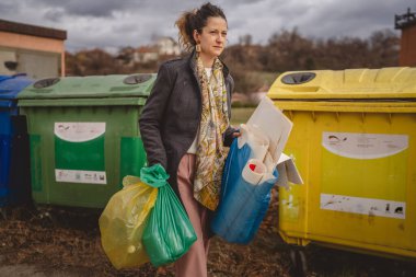 Bir yetişkin kadın cam, kağıt ve plastik sıfır atık konsepti için geri dönüşüm kutularının önünde yeşil sarı ve mavi poşetlerle duruyor.