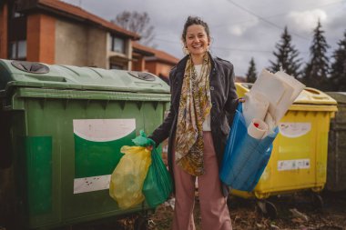 Bir yetişkin kadın cam, kağıt ve plastik sıfır atık konsepti için geri dönüşüm kutularının önünde yeşil sarı ve mavi poşetlerle duruyor.