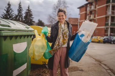 Bir yetişkin kadın cam, kağıt ve plastik sıfır atık konsepti için geri dönüşüm kutularının önünde yeşil sarı ve mavi poşetlerle duruyor.