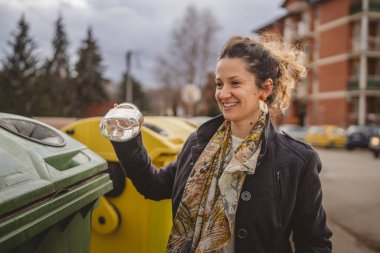 Bir yetişkin kadın cam atık ve yeşil poşetlerle geri dönüşüm kaplarının önünde duruyor ve sıfır atık eko kavramını yeniden kullanmak için şişe kavanozu fırlatıyor.