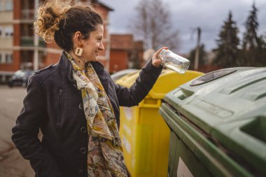 Bir yetişkin kadın cam atık ve yeşil poşetlerle geri dönüşüm kaplarının önünde duruyor ve sıfır atık eko kavramını yeniden kullanmak için şişe kavanozu fırlatıyor.