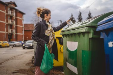Bir yetişkin kadın cam atık ve yeşil poşetlerle geri dönüşüm kaplarının önünde duruyor ve sıfır atık eko kavramını yeniden kullanmak için şişe kavanozu fırlatıyor.