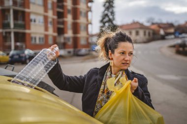 Bir yetişkin kadın, plastik atık ve sarı poşetlerle geri dönüşüm kutularının önünde duruyor ve sıfır atık eko kavramını yeniden kullanmak için şişe fırlatıyor.