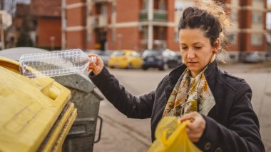Bir yetişkin kadın, plastik atık ve sarı poşetlerle geri dönüşüm kutularının önünde duruyor ve sıfır atık eko kavramını yeniden kullanmak için şişe fırlatıyor.